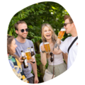 Vier Personen stoßen mit Biergläsern und Bierflaschen an, umgeben von grünen Bäumen im Freien