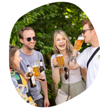 Vier Personen stoßen mit Biergläsern und Bierflaschen an, umgeben von grünen Bäumen im Freien