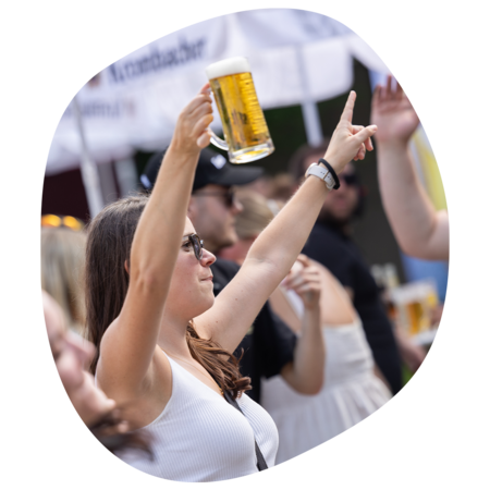 Frau in weißem Top hält Bierkrug hoch und zeigt mit der anderen Hand nach oben