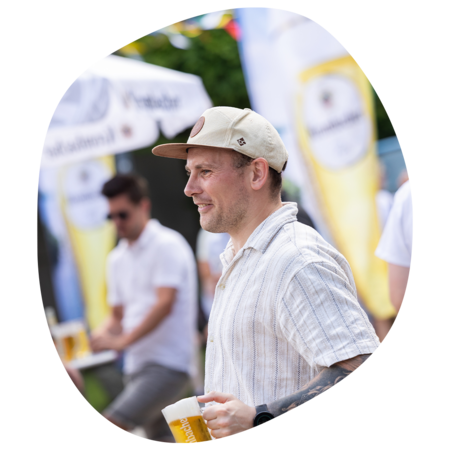 Mann mit hellbeiger Kappe und gestreiftem Hemd hält ein Glas Bier auf dem Krombacher Sommerfest