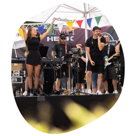 Band spielt auf Bühne unter Zelt mit bunten Wimpeln, Sängerin hebt Arme, Musiker mit Gitarre und Keyboard