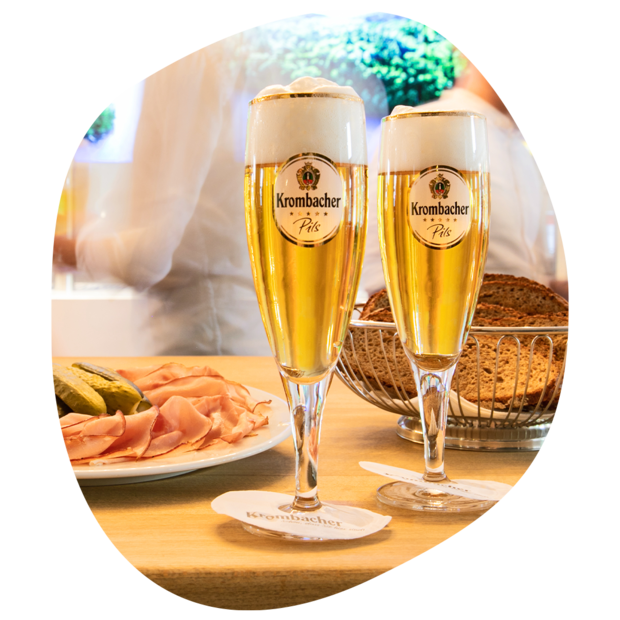 Zwei Gläser Krombacher Pils mit Schaum auf einem Holztisch, daneben Teller mit Schinken und Gurken sowie ein Brotkorb mit dunklem Brot.