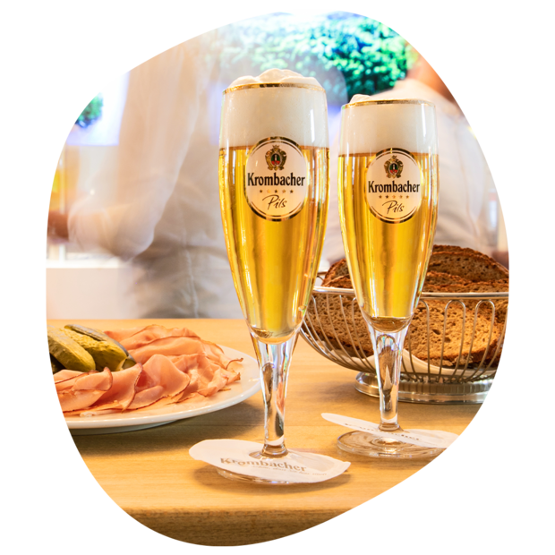 Zwei Gläser Krombacher Pils mit Schaum auf einem Holztisch, daneben Teller mit Schinken und Gurken sowie ein Brotkorb mit dunklem Brot.