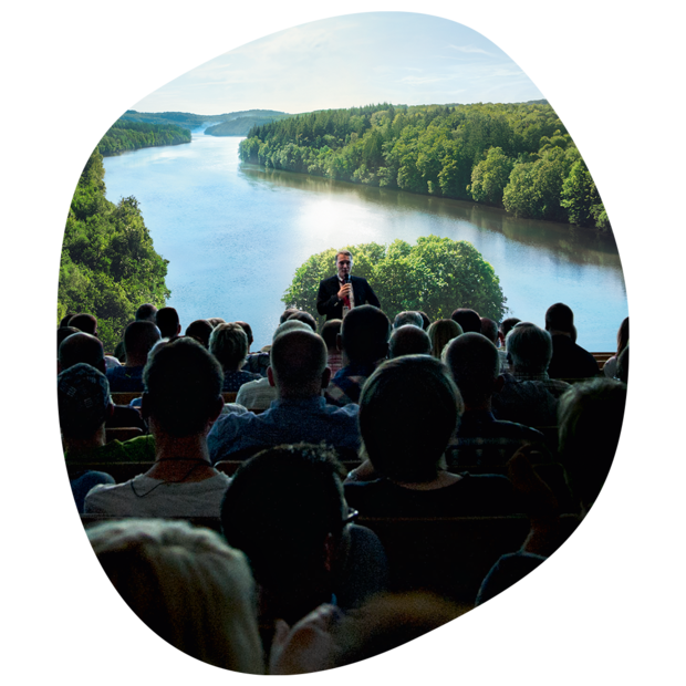 Publikum sitzt in einem 180-Grad-Panoramakino blickt auf einen See, umgeben von dicht bewaldeten Hügeln und blauem Himmel.