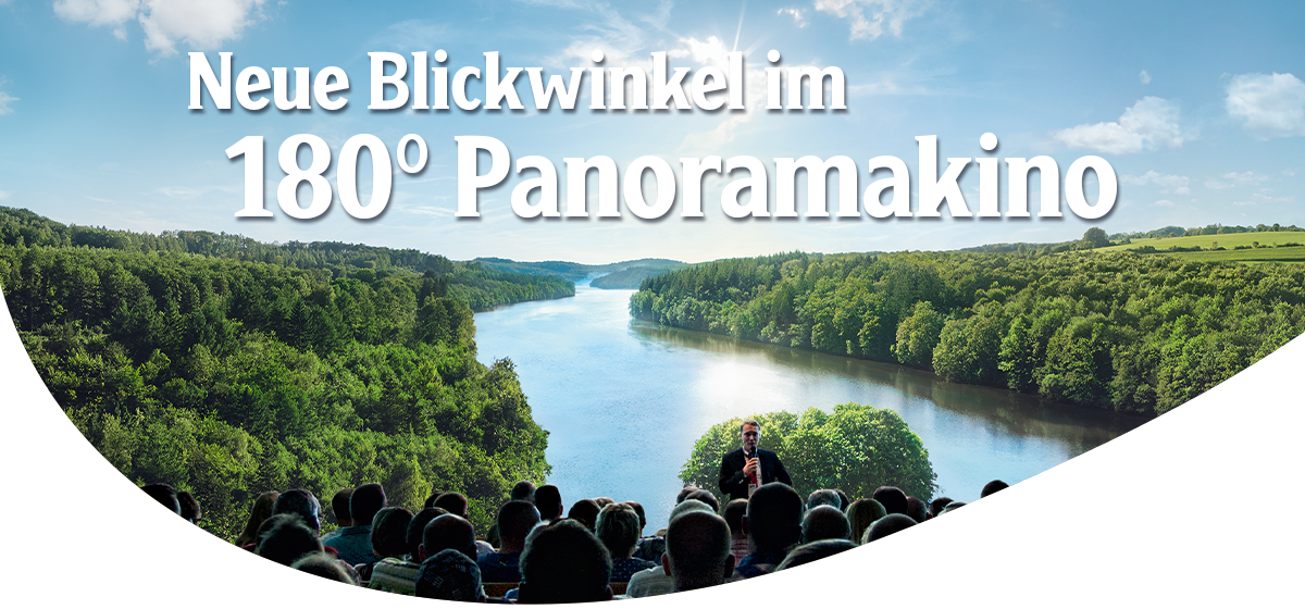 Publikum sitzt in einem 180-Grad-Panoramakino blickt auf einen See, umgeben von dicht bewaldeten Hügeln und blauem Himmel, Headline 'Neue Blickwinkel im 180° Panoramakino'.