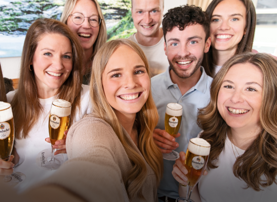 Gruppe von acht lächelnden jungen Erwachsenen, die gemeinsam ein Selfie machen und Krombacher Pils in Gläsern in der Hand halten. 