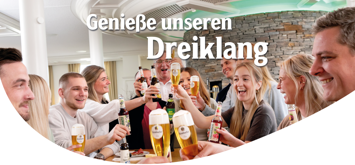 Eine Gruppe von gut gelaunten Menschen sitzen zusammen und erheben ihre Gläser und Flaschen zum Anstoßen. Headline: Genieße unseren Dreiklang.
