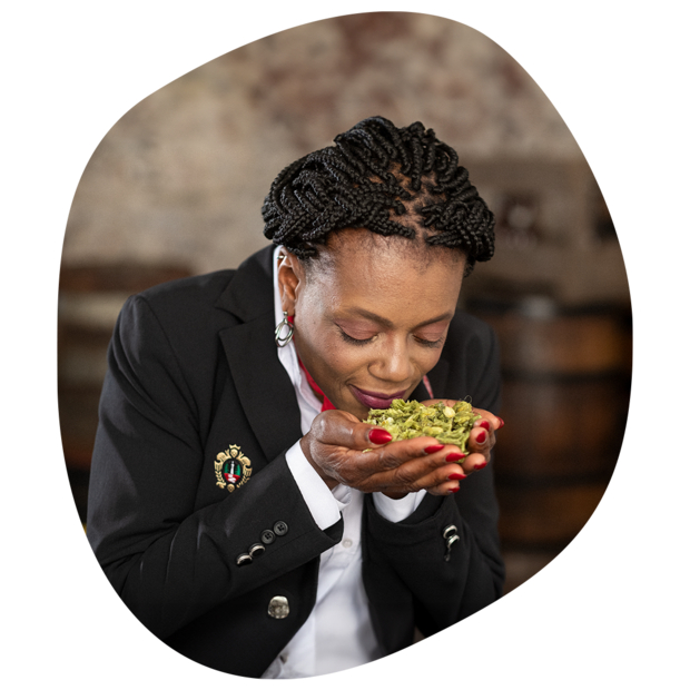 Frau mit geflochtenem schwarzen Haar und schwarzem Blazer hält frischen Hopfen in den Händen und riecht daran.