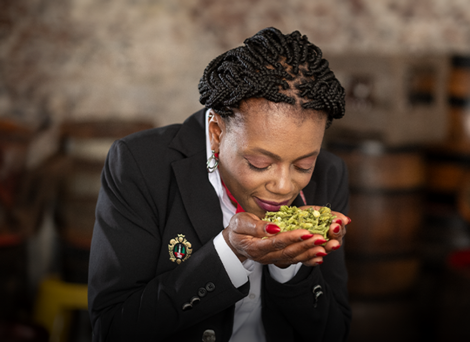 Frau mit schwarzen geflochtenen Haaren hält ein paar Hopfen in den Händen und riecht daran.