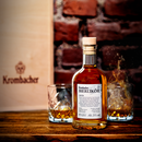 Flasche Krombacher Bierlikör vor Holzkiste mit Krombacher-Logo und zwei Gläser mit Likör auf einem Holztisch.