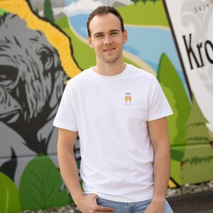 Ein Mann in einem weißen T-Shirt mit zwei anstoßenden Krombacher Gläsern auf der Brust, vor einer bunten Wand.