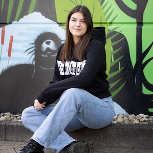 Person sitzt auf dem Boden vor einer Wand mit Bären-Graffiti, trägt schwarze Krombacher Sweatshirtjacke und blaue Jeans.