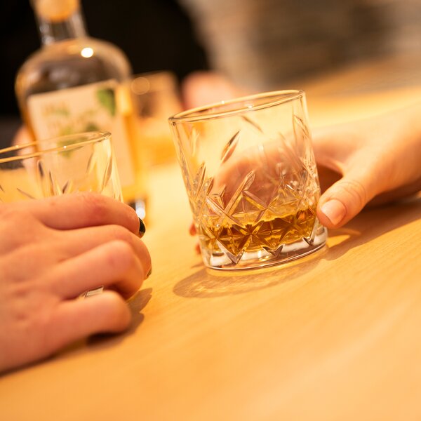 Zwei Hände halten Kristallgläser mit Bierbrand auf einem Holztisch.