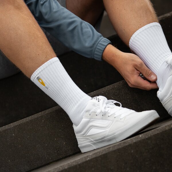 Person zieht weiße Socken mit kleinem gelbem Pokal-Logo an, trägt weiße Sportschuhe und graue Sporthose.