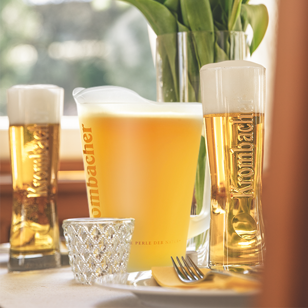 Krombacher Pils im Pitcher und zwei Gläsern auf einem gedeckten Tisch.