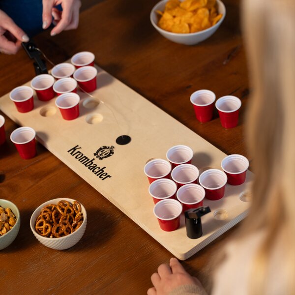 Mini Beerpong-Spielbrett mit roten Bechern und Krombacher-Logo, umgeben von Snacks wie Brezeln und Chips, Hände beim Spielen.