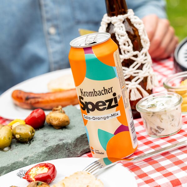 Krombacher Spezi Cola-Orange Dose auf einem Tisch mit Grillgerichten, Snacks und Dips.