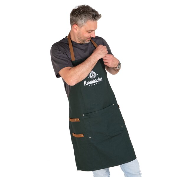 Mann trägt dunkel grüne Krombacher-Grillschürze mit braunen Trägern und Taschen über schwarzem T-Shirt und hellen Jeans