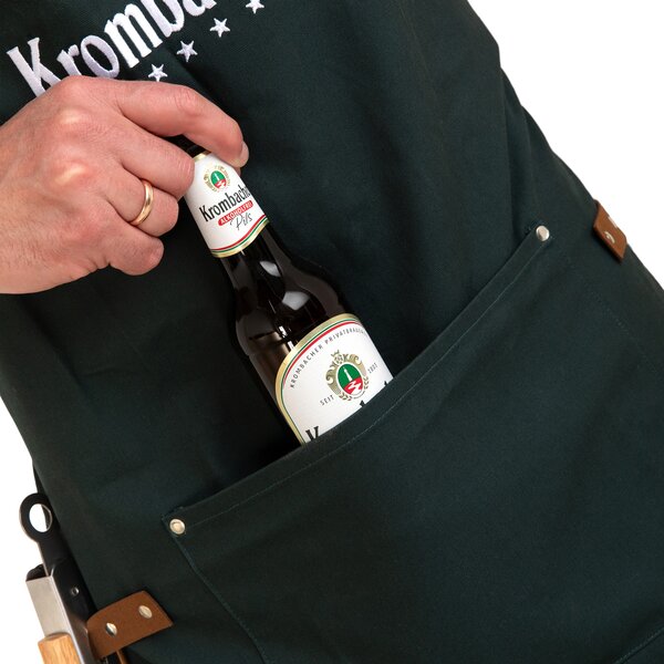 Hand steckt eine Flasche Krombacher Pils in die Tasche einer dunklen Grillschürze mit sichtbarem Krombacher-Logo