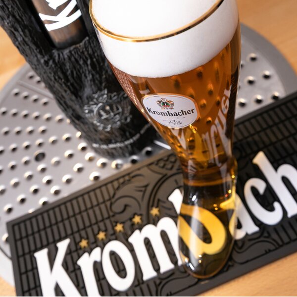 Frisch gezapftes Glas Bier in Stiefelform mit Krombacher-Logo, stehend auf einer schwarzen Tropfmatte mit Krombacher-Schriftzug.