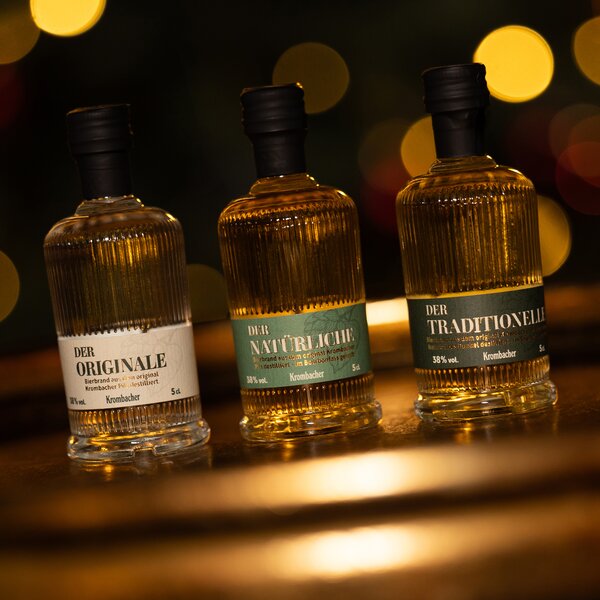 5555_krombachershop_Probierbrand-Set_Mood_001 Drei kleine Krombacher Spirituosenflaschen mit schwarzen Verschlüssen, beschriftet mit 'Der Originale', 'Der Natürliche' und 'Der Traditionelle', vor unscharfem Lichthintergrund.