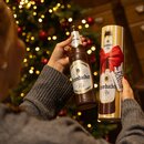 Person hält eine braune Krombacher Pils Flasche aus Schokolade und eine Geschenkverpackung mit roter Schleife und Aufschrift 'Schokoladen Flasche'.