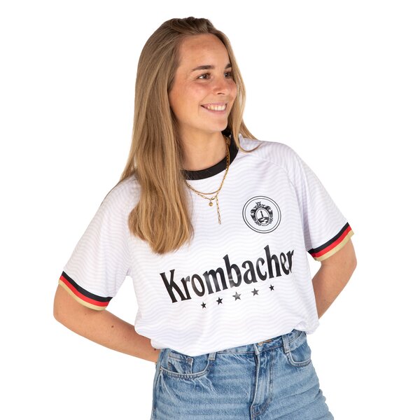 Frau trägt weißes Trikot mit schwarzem Kragen, Krombacher Logo und schwarz-rot-goldenen Ärmelbündchen mit blauer Jeans.