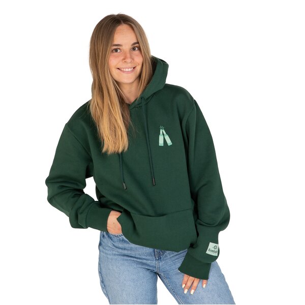 Frau trägt dunkelgrünen Hoodie mit kleinem Flaschendruck auf der Brust und Jeans, eine Hand in der Hosentasche