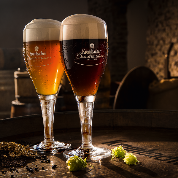 Zwei Krombacher Brautradition Biergläser, eines bernsteinfarben, das andere dunkel, auf Holztisch mit Hopfen und Malz.