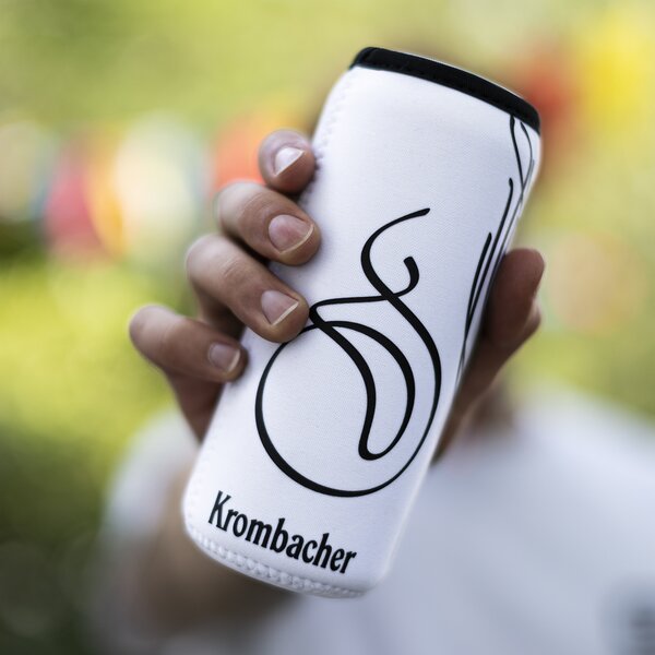 Hand hält weißen Dosenkühler mit schwarzer Krombacher-Logo-Schrift und geschwungenem Design.